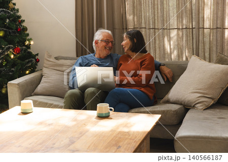 Senior couple sitting on sofa using silver laptop while holding mugs by Christmas tree with pillows Senior couple sitting on sofa using silver laptop while holding mugs by Christmas tree with pillows 140566187