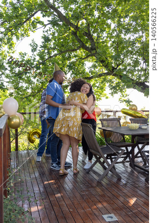 Diverse friends hugging under pastel pink, gold balloons on backyard deck near snack bowl 140566225