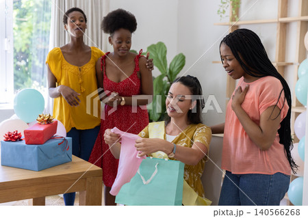 Diverse female friends gathering in home living room and opening pink baby onesie from gift bag Diverse female friends gathering in home living room and opening pink baby onesie from gift bag 140566268