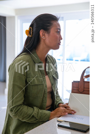 Asian woman sitting at kitchen counter writing in notebook with smartphone by tote bag Asian woman sitting at kitchen counter writing in notebook with smartphone by tote bag 140566311