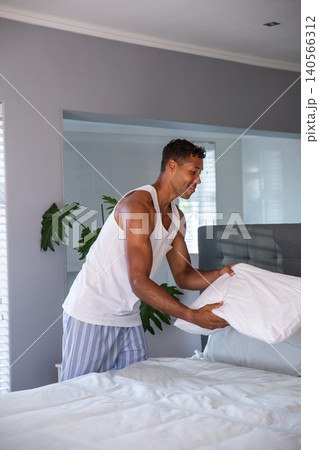 African american mid adult man standing and straightening pillow on white bedding by headboard African american mid adult man standing and straightening pillow on white bedding by headboard 140566312