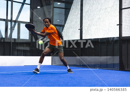 African American male athlete swinging paddle racket at neon green ball on blue padel court 140566385