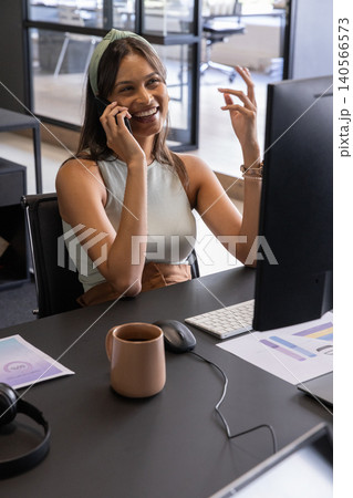 Woman sitting at office desk holding smartphone, gesturing near keyboard, charts, headphones 140566573