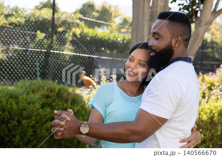 Diverse couple dancing holding hands in garden under trees near chain-link fence wearing wristwatch Diverse couple dancing holding hands in garden under trees near chain-link fence wearing wristwatch 140566670