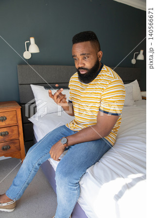 African American man sitting on edge of bed in bedroom checking smartphone, glancing at wristwatch African American man sitting on edge of bed in bedroom checking smartphone, glancing at wristwatch 140566671