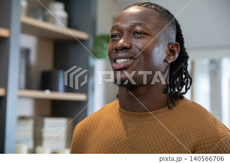 Smiling african american man in brown sweatshirt looking upward in home office with bookshelf Smiling african american man in brown sweatshirt looking upward in home office with bookshelf 140566706