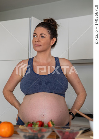 Pregnant woman in sports bra standing at kitchen counter surveying fruit bowls and cutting board Pregnant woman in sports bra standing at kitchen counter surveying fruit bowls and cutting board 140566719