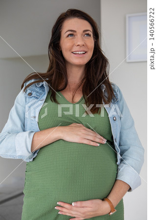 Pregnant woman cradling bump in green dress wearing denim jacket in hallway with wall art Pregnant woman cradling bump in green dress wearing denim jacket in hallway with wall art 140566722
