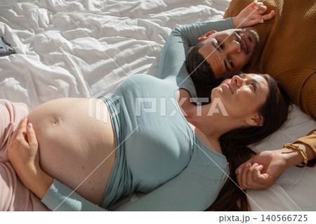 Diverse couple lying together on white bed sheets in softly lit bedroom holding hands near pillow 140566725