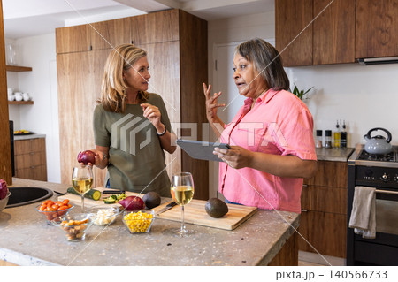 Diverse middle-aged female friends cooking at kitchen countertop using tablet while slicing onion Diverse middle-aged female friends cooking at kitchen countertop using tablet while slicing onion 140566733