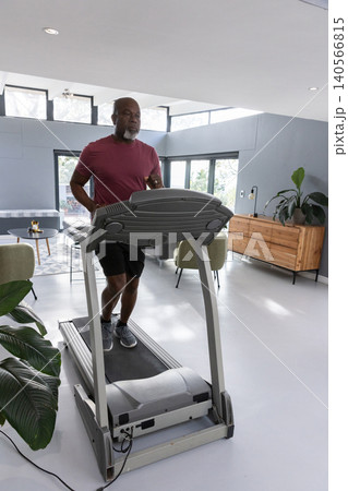 Senior african american man jogging on treadmill in living room near plants and chairs Senior african american man jogging on treadmill in living room near plants and chairs 140566815