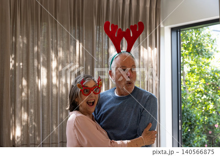 Senior couple hugging and showing surprise at home with red reindeer glasses and antler headband 140566875