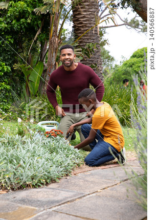 African american father and son kneeling in yard digging, trowel in flowerbed with watering can African american father and son kneeling in yard digging, trowel in flowerbed with watering can 140566887