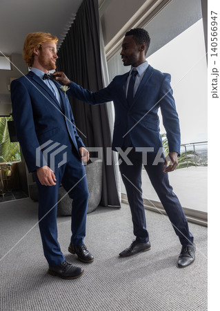 Groom and groomsman adjusting bow tie and boutonniere in suite by balcony in navy-blue suits 140566947