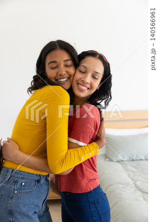 Diverse mom and teenage daughter hugging wearing yellow top and red t-shirt in bedroom Diverse mom and teenage daughter hugging wearing yellow top and red t-shirt in bedroom 140566961