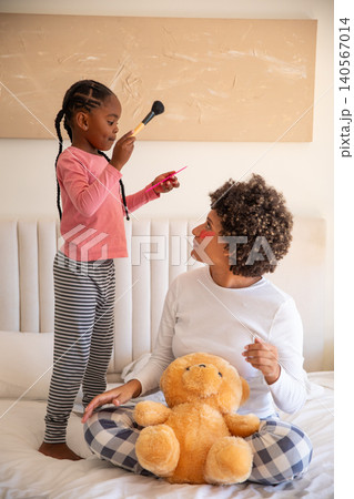 Diverse mother with daughter playing makeup on bed in bedroom with makeup brush and lip gloss Diverse mother with daughter playing makeup on bed in bedroom with makeup brush and lip gloss 140567014