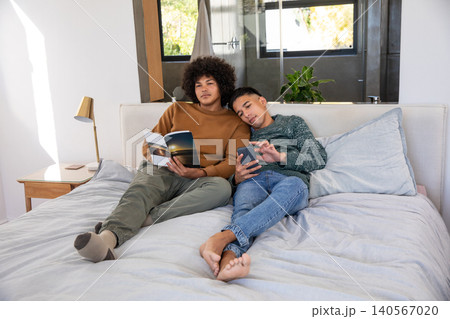 Male couple sitting on bed reading book and scrolling smartphone in modern bedroom with lamp Male couple sitting on bed reading book and scrolling smartphone in modern bedroom with lamp 140567020