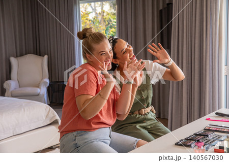 Female friends waving at camera sitting at table in bedroom with eyeshadow palettes 140567030