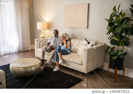 Diverse senior couple sitting on corduroy sofa in living room with coffee table and potted plant Diverse senior couple sitting on corduroy sofa in living room with coffee table and potted plant 140567032