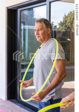 Senior man holding neon hula hoop on patio by sliding glass door in T-shirt and shorts 140567374