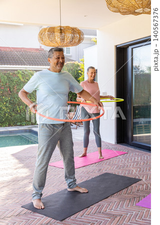 Mature couple exercising using orange and yellow hula hoops on covered patio beside pool, on mats Mature couple exercising using orange and yellow hula hoops on covered patio beside pool, on mats 140567376