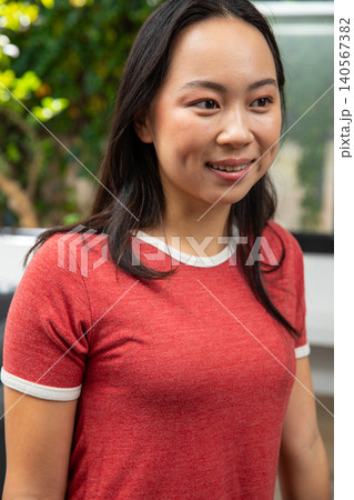 Adult Asian woman posing by window smiling looking right wearing red tee white trim necklace plants 140567382
