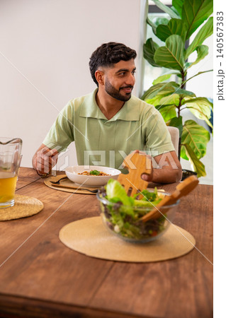 Adult Indian man wearing light green polo sitting at home table with salad bowl, looking right Adult Indian man wearing light green polo sitting at home table with salad bowl, looking right 140567383