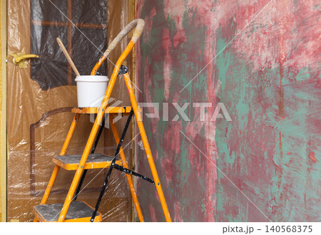 Home renovation, interior painting project with yellow step ladder, paint bucket, mixing stick beside protected wooden door. Colorful textured wall with pink and turquoise paint DIY decoration concept Home renovation, interior painting project with yellow step ladder, paint bucket, mixing stick beside protected wooden door. Colorful textured wall with pink and turquoise paint DIY decoration concept 140568375