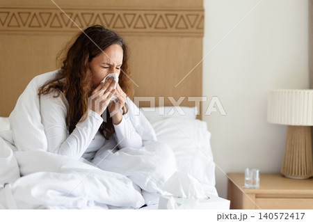 Woman sneezing into tissue while resting in bed, showing cold symptoms Woman sneezing into tissue while resting in bed, showing cold symptoms 140572417