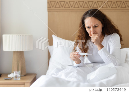 Sick woman coughing while checking thermometer in bed, illustrating respiratory illness, Sick woman coughing while checking thermometer in bed, illustrating respiratory illness, 140572449