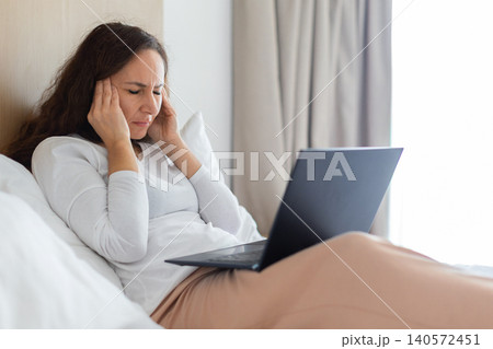 Woman resting in bed with laptop while suffering from headache Woman resting in bed with laptop while suffering from headache 140572451