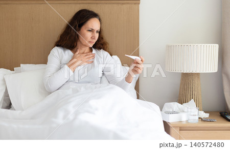 Concerned woman checking thermometer while sitting in bed, monitoring fever symptoms 140572480