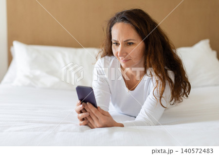 Relaxed adult woman lying on bed and using smartphone, browsing online content 140572483