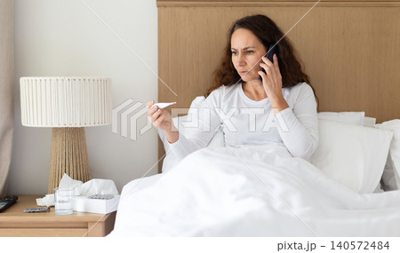 Woman talking on smartphone while checking thermometer in bed Woman talking on smartphone while checking thermometer in bed 140572484