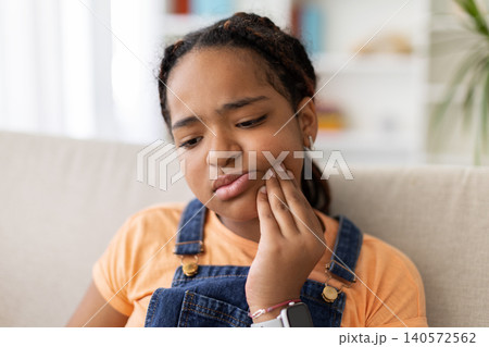 Black teenage girl touching cheek with concerned expression at home, suffering toothache Black teenage girl touching cheek with concerned expression at home, suffering toothache 140572562