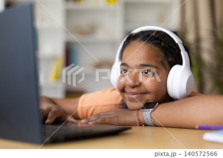 Black teenage girl smiling while using laptop and wearing headphones at home Black teenage girl smiling while using laptop and wearing headphones at home 140572566