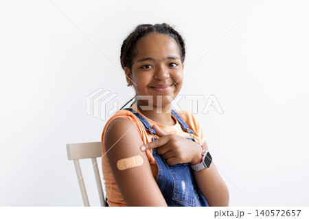 Black teenage girl pointing to bandage on her upper arm after vaccination Black teenage girl pointing to bandage on her upper arm after vaccination 140572657