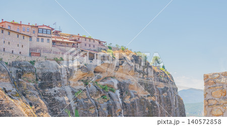 Wide panoramic view of the traditional stone buildings of Great Meteoron monastery on a giant rock pillar, showcasing long steel transport cables stretching over a canyon Wide panoramic view of the traditional stone buildings of Great Meteoron monastery on a giant rock pillar, showcasing long steel transport cables stretching over a canyon 140572858