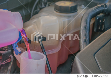 Bright pink washer fluid pouring from a plastic canister into the vertical reservoir tank neck under an open car hood during regular automotive maintenance service Bright pink washer fluid pouring from a plastic canister into the vertical reservoir tank neck under an open car hood during regular automotive maintenance service 140572868