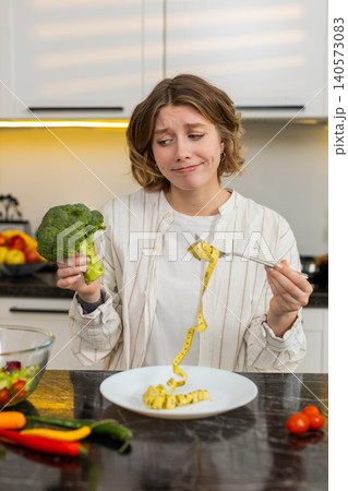 Young woman feeling upset with diet choice in home kitchen holding broccoli during slimming routine Young woman feeling upset with diet choice in home kitchen holding broccoli during slimming routine 140573083