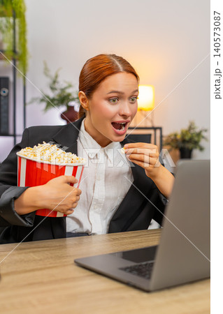 Young businesswoman enjoys popcorn watching movie on laptop during lunch break smiling at fun scene Young businesswoman enjoys popcorn watching movie on laptop during lunch break smiling at fun scene 140573087