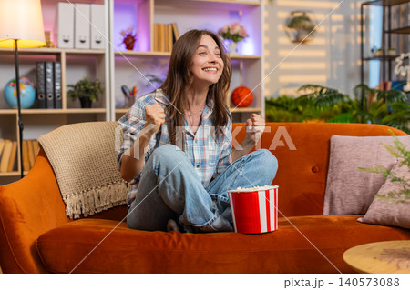 Young woman on home sofa cheering with popcorn while watching sports and celebrating team success Young woman on home sofa cheering with popcorn while watching sports and celebrating team success 140573088