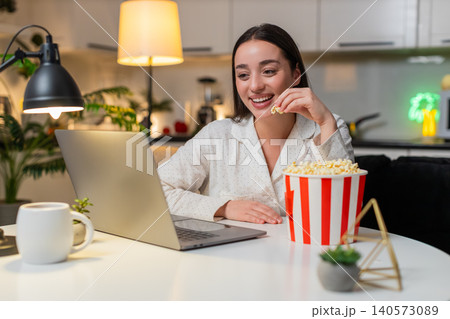 Young woman watches movie trailer on laptop with popcorn laughs comments reacts evening fun with joy Young woman watches movie trailer on laptop with popcorn laughs comments reacts evening fun with joy 140573089