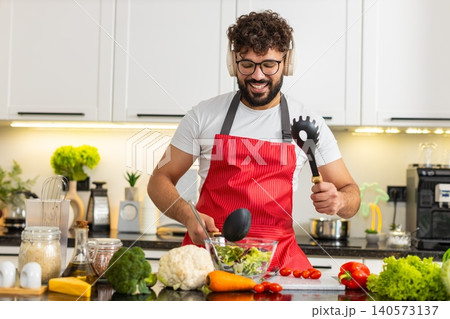 Arabian young adult man dancing in kitchen cooking salad listening to music in wireless headphones Arabian young adult man dancing in kitchen cooking salad listening to music in wireless headphones 140573137