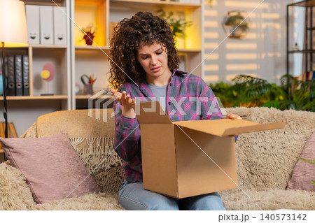 Young woman on home sofa opening delivery box then feeling upset over wrong item from online store 140573142
