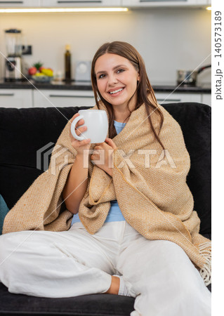 Young woman drinks herbal tea holds mug takes slow sip wraps in blanket relaxes and enjoys warm rest Young woman drinks herbal tea holds mug takes slow sip wraps in blanket relaxes and enjoys warm rest 140573189