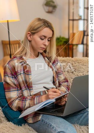 Adult woman writing financial notes in notebook while looking at laptop screen at home with focus 140573197