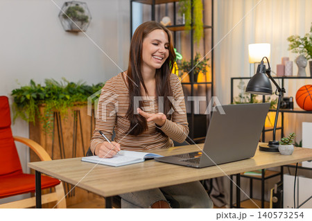 Young woman in headphones on video call gestures and writes notes in notebook while focused on work Young woman in headphones on video call gestures and writes notes in notebook while focused on work 140573254