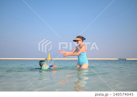 Asian woman in swimsuit plays with multiracial son in shallow water at beach Asian woman in swimsuit plays with multiracial son in shallow water at beach 140574045