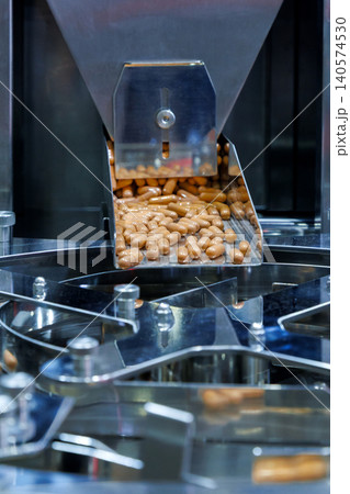brown medicine capsules or dietary supplements falling from a metal chute into a rotary sorting machine. Pharmaceutical manufacturing and industrial mass production packaging process. brown medicine capsules or dietary supplements falling from a metal chute into a rotary sorting machine. Pharmaceutical manufacturing and industrial mass production packaging process. 140574530